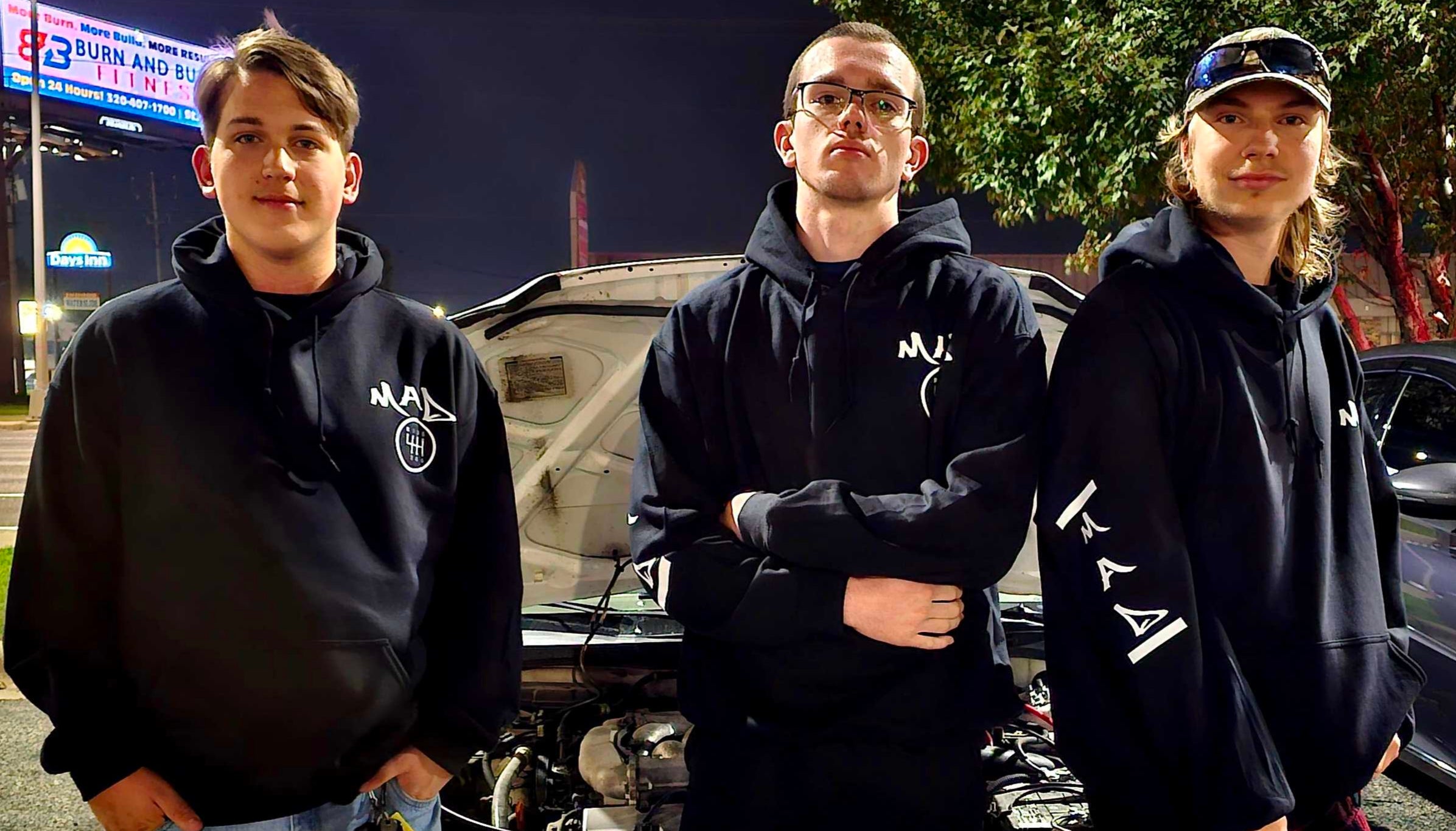 Three individuals standing in front of a car with an open hood at night.